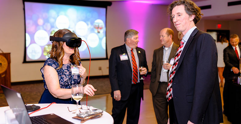 A woman in a blue dress uses a VR headset at a formal event while others in suits converse nearby.