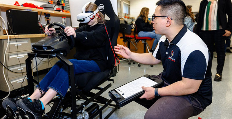 A person using a racing simulator with a VR headset while another person crouches beside them with a clipboard in a workshop setting.
