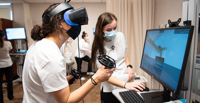Two individuals in white uniforms use virtual reality equipment in a medical simulation room.